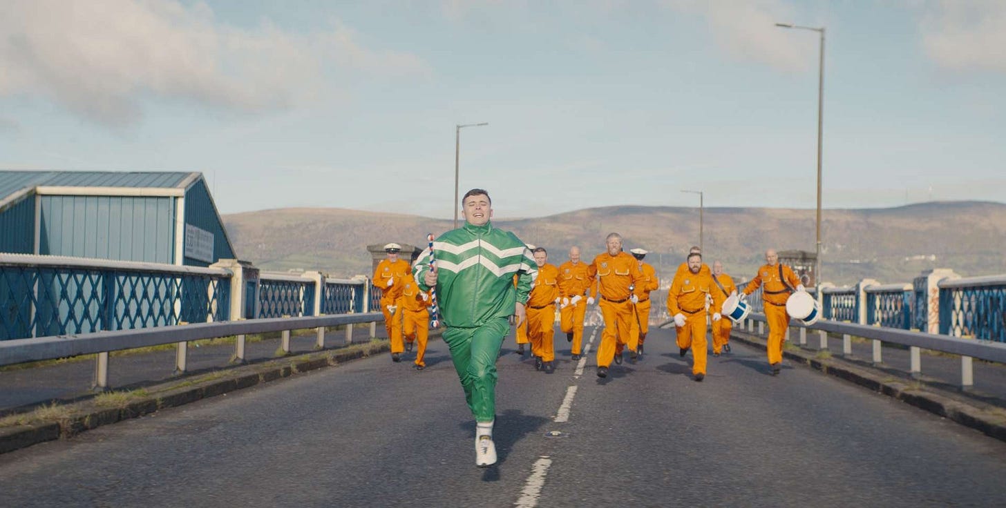 Kneecap's Mo Chara running towards the camera wearing a green tracksuit. He is pursued by a group of orange-clad protestants.