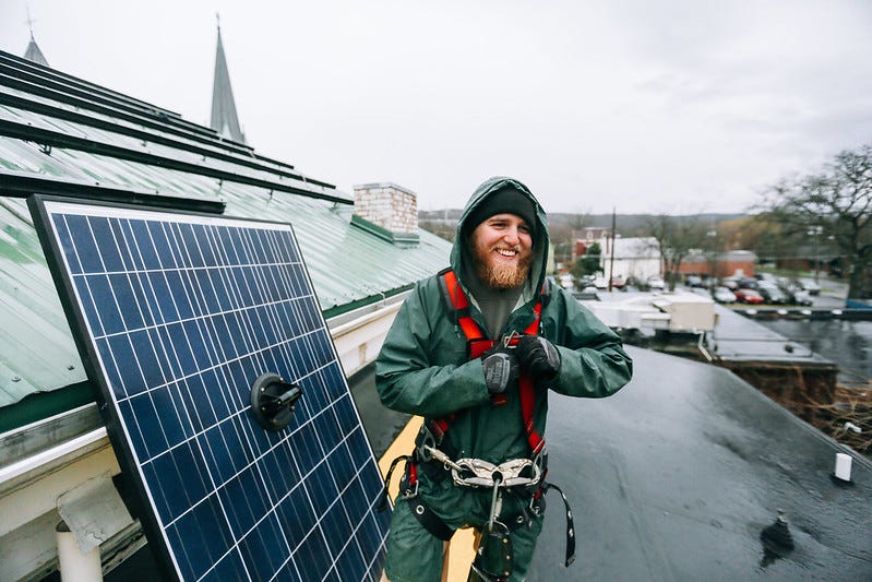 A solar engineer clad all in green installs panels on a roof in the rain.