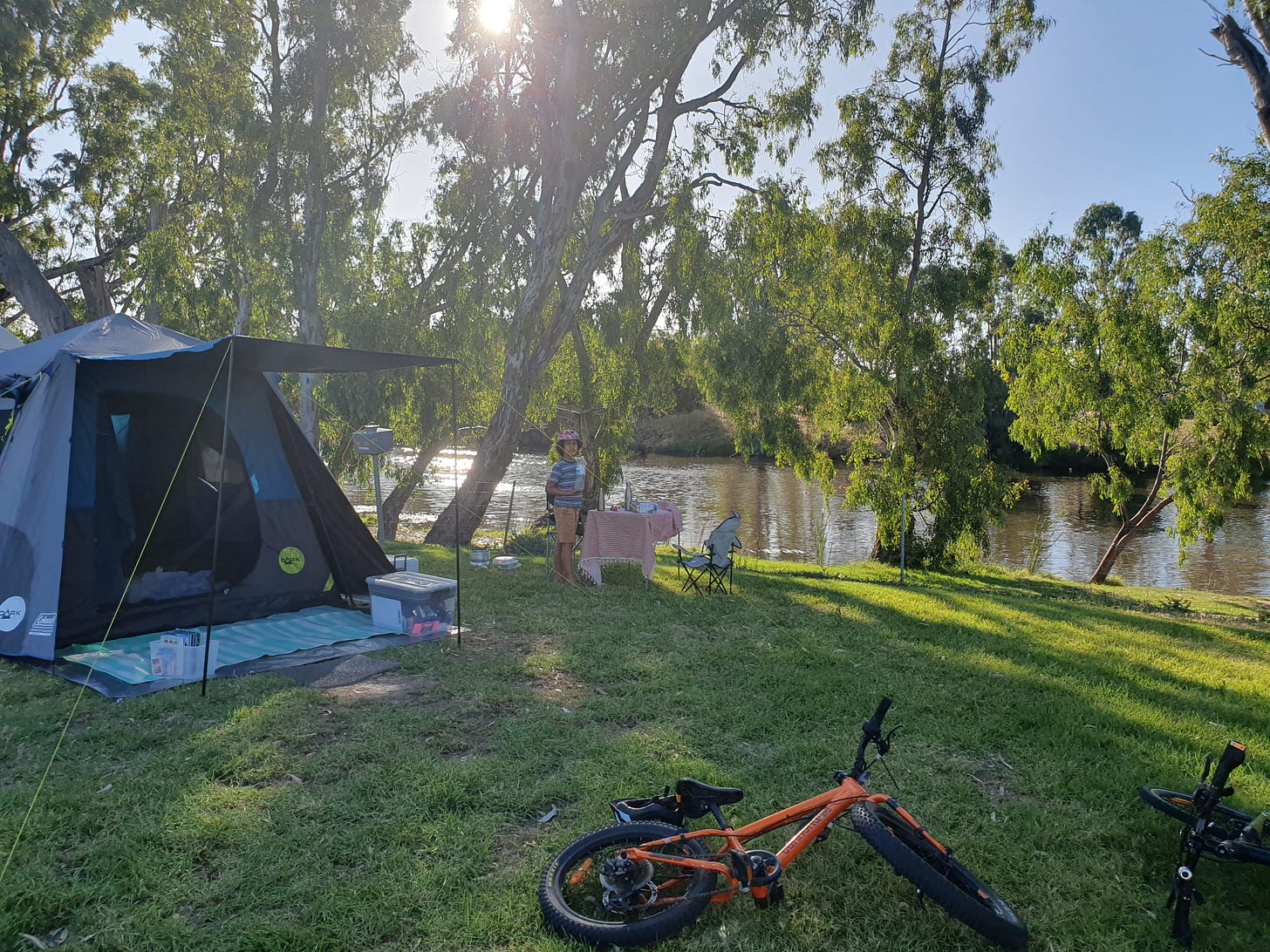 A tent by the river. In the foreground there are bikes on the grass and in the background a child beside a table and chair.