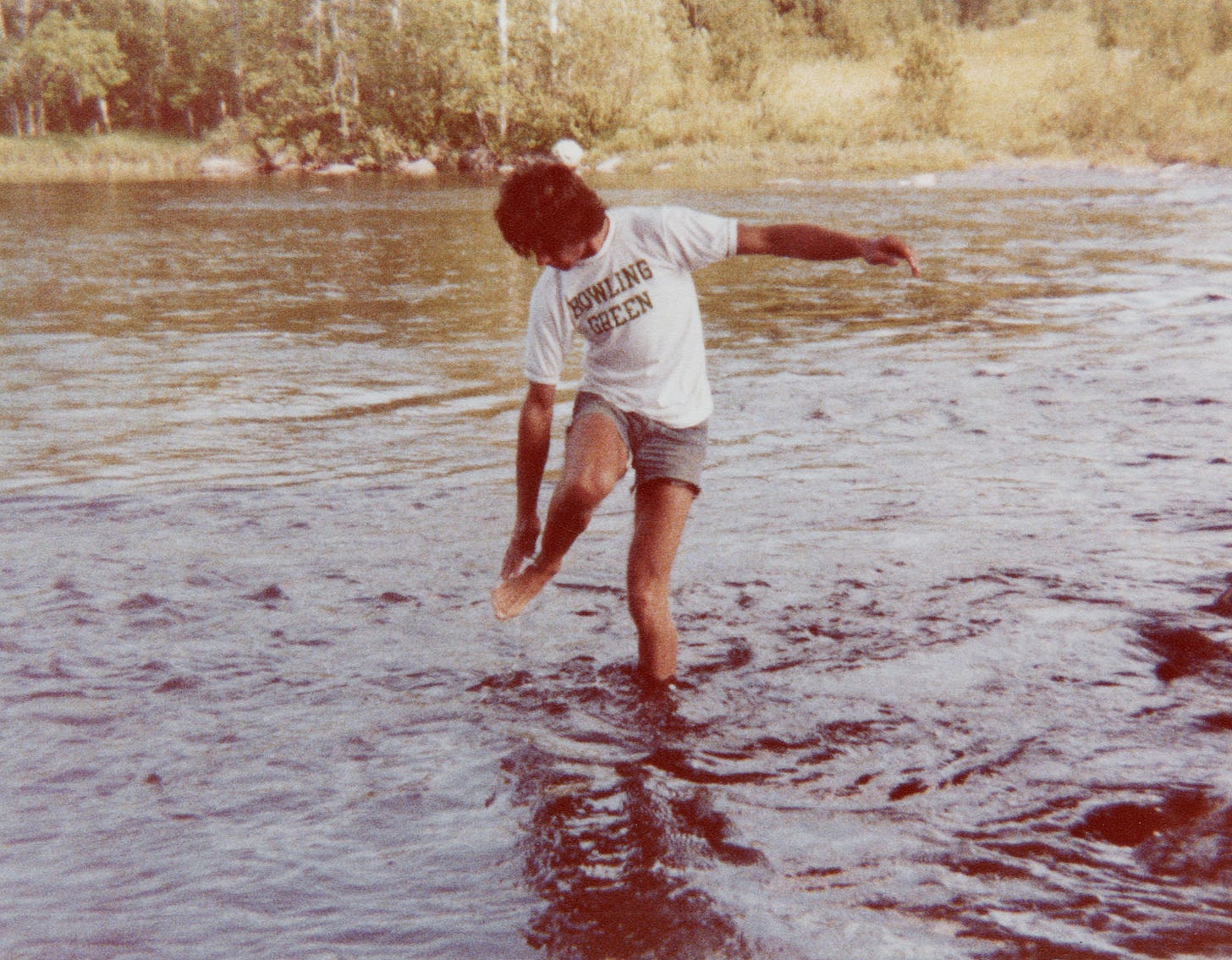 A picture of me wading in shin-deep water of a river somewhere between Minneapolis and the north shore of Lake Superior. I'm wearing a t-shirt with BOWLING GREEN in two lines across my chest, and I'm reaching down to touch my right foot which is raised about a foot above the water. I must have stepped on something. My left arm is almost perpendicular to my body as I balance myself. I'm looking down at my right foot. Only a bit of my face is showing. I'm wearing cut-off blue jean shorts which show probably two-thirds of my thigh.