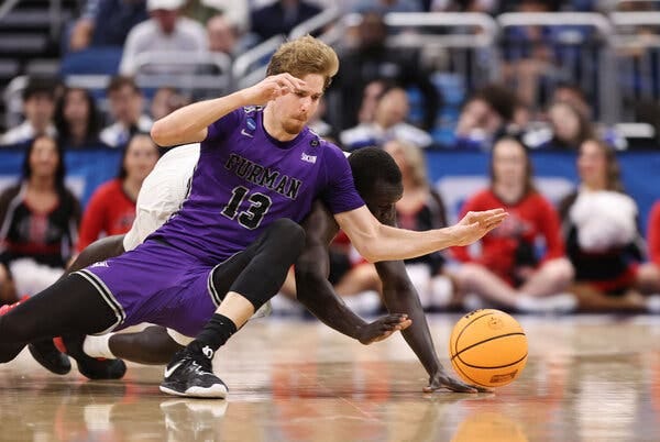 Garrett Hien, in a No. 13 purple Furman jersey, reaches for the ball on the floor while falling into Aguek Arop of San Diego State, also diving.