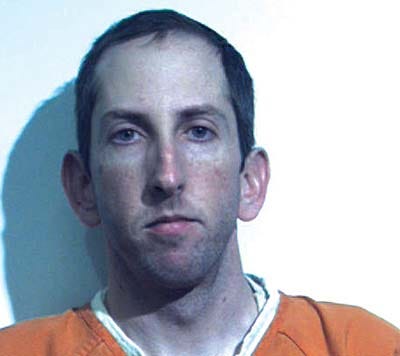 A man in an orange prison shirt facing the camera against a white wall