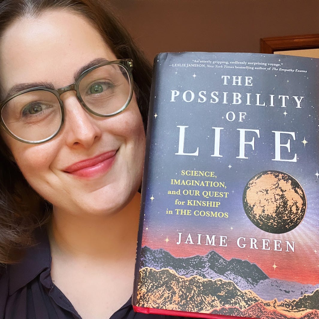 me, a white woman with brown hair and glasses, smiling and holding up a copy of my book