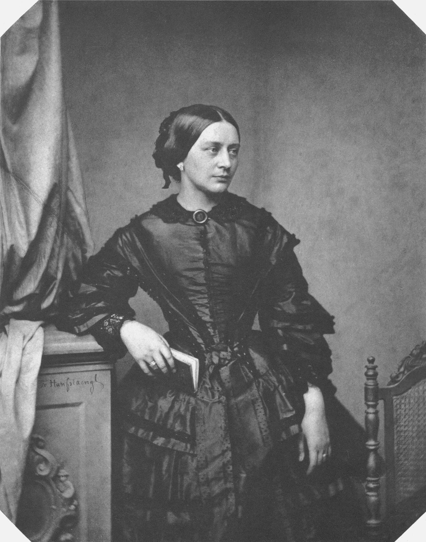 Black and white photo of Clara Schumann in her 30s, wearing an ornate black gown and holding a leatherbound book in her right hand as she poses against a chest of drawers