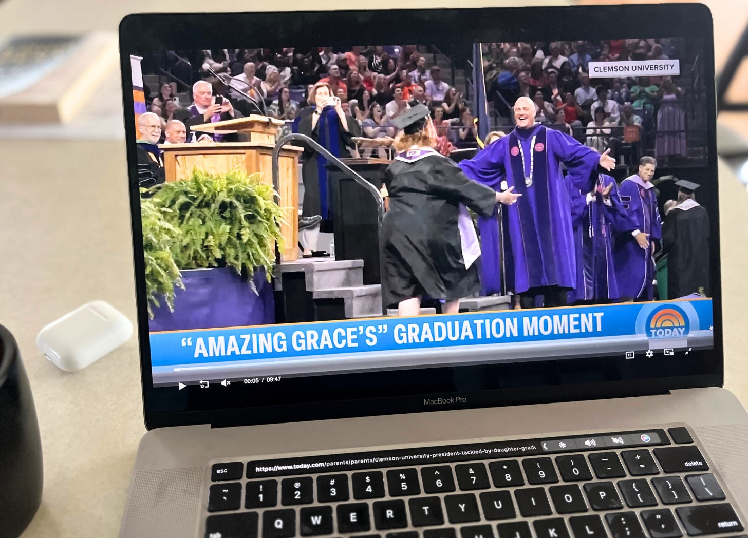 a laptop screen displays a graduation ceremony with a graduate in a cap and gown hugging an older man in academic regalia, with the text “Clemson University” and “GRACING GRACE'S GRADUATION MOMENT” on the screen. a laptop screen displays a graduation ceremony with a graduate in a cap and gown hugging an older man in academic regalia, with the text “Clemson University” and “GRACING GRACE'S GRADUATION MOMENT” on the screen.