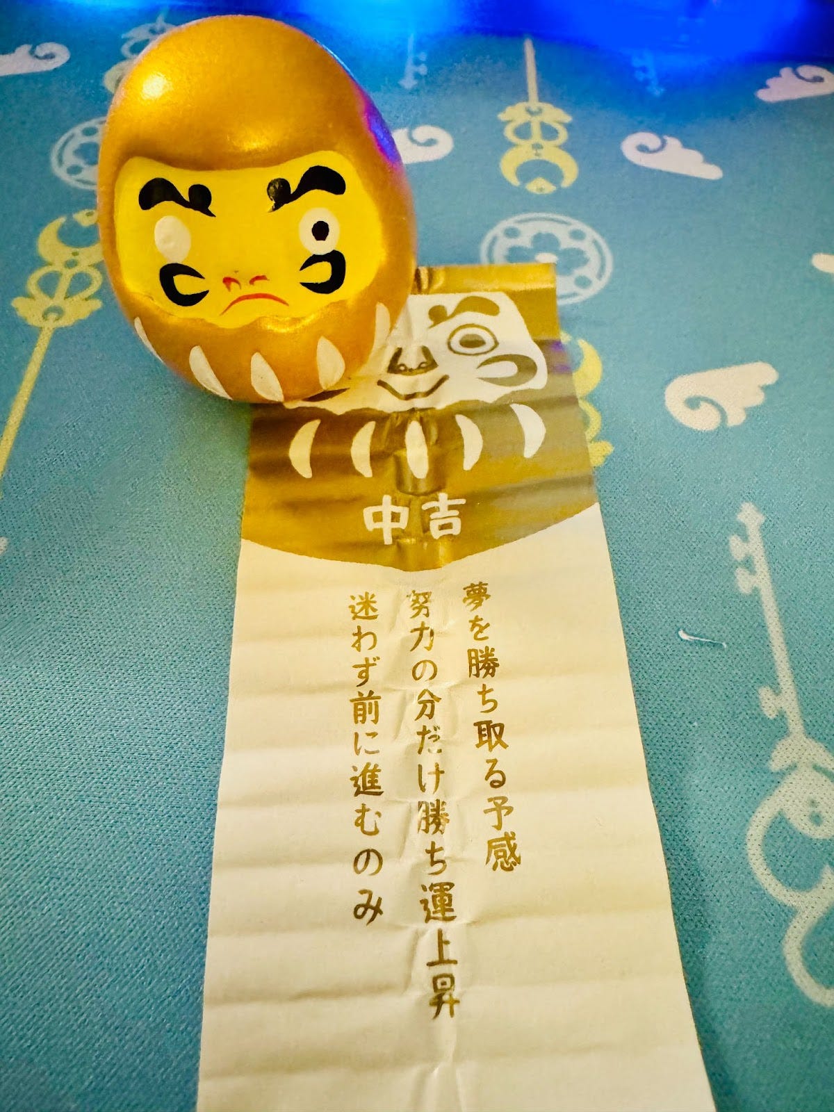 A golden Daruma doll (a Japanese symbol for perseverence and good luck) with its left eye painted, and a fortune underneath reading "middle blessing".
