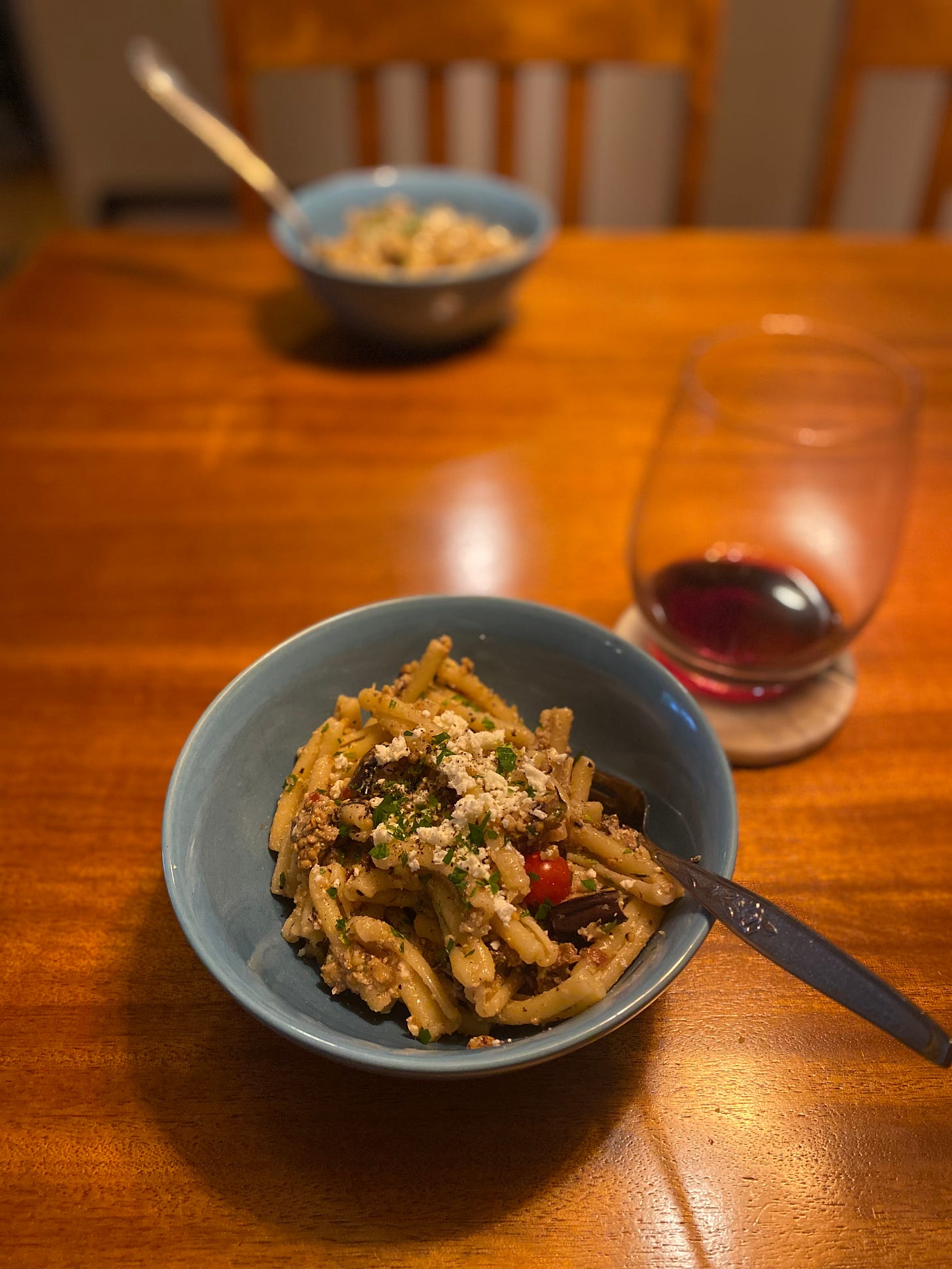 Two blue bowls across from each other on a table, each with pasta salad made with casarecce. The salad has pieces of eggplant and small red tomatoes throughout, and the top is covered with ground pepper and crumbled feta. A half-finished glass of red wine rests on the coaster behind the nearest bowl.