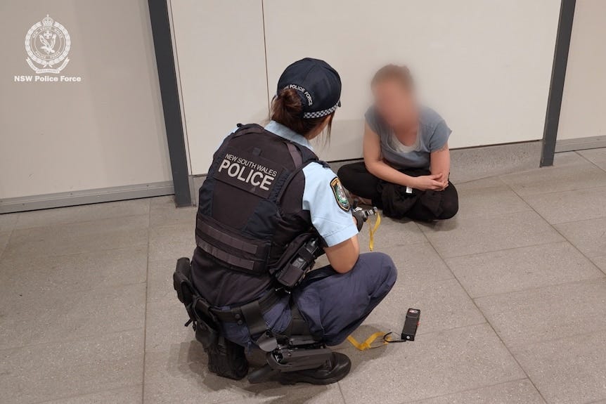 A female police officer crouches by a woman with a slingshot and taser next to her.