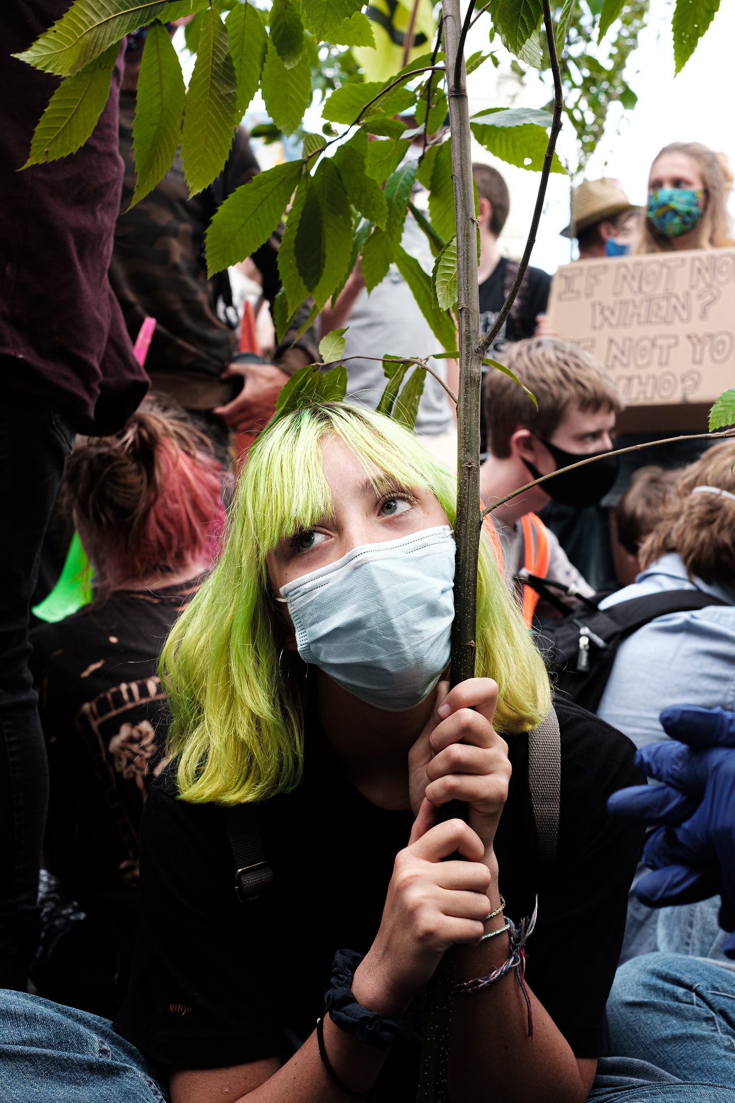 a white woman with yellow-green hair, wearing a face mask, holding up some kind of plant at a protest.