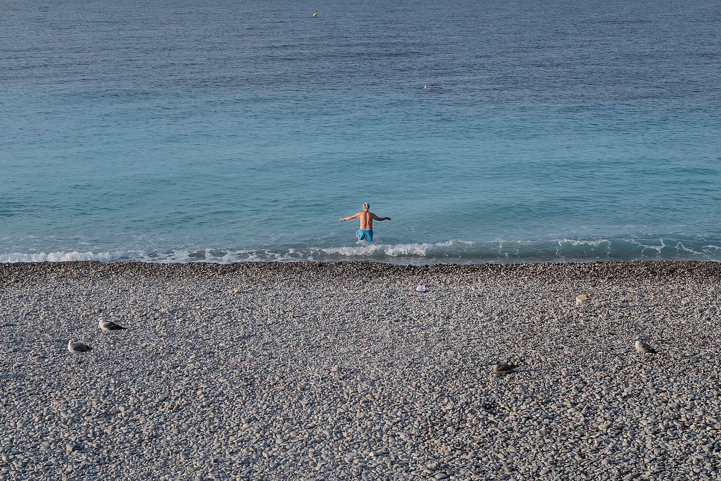 Solitary Swimmer in the Mediterranean – A peaceful beach scene in Nice, where a lone swimmer stands waist-deep in the clear blue water, framed by the pebbled shore.