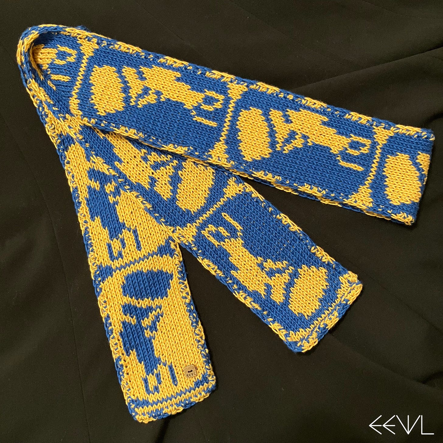 Blue and yellow/gold double-sided hand-knit scarf featuring the portrait bust of Queen Nefertiti in profile. Scarf is folded on a black fabric background.