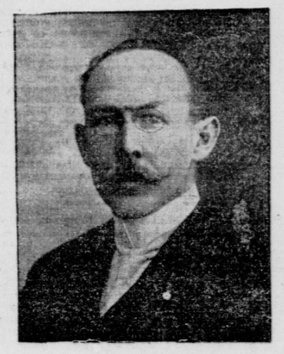 A photographic portrait of a white man with a receding hairline, a moustache and little round glasses. He wears a high collar and a suit jacket. His ears are somewhat prominent.