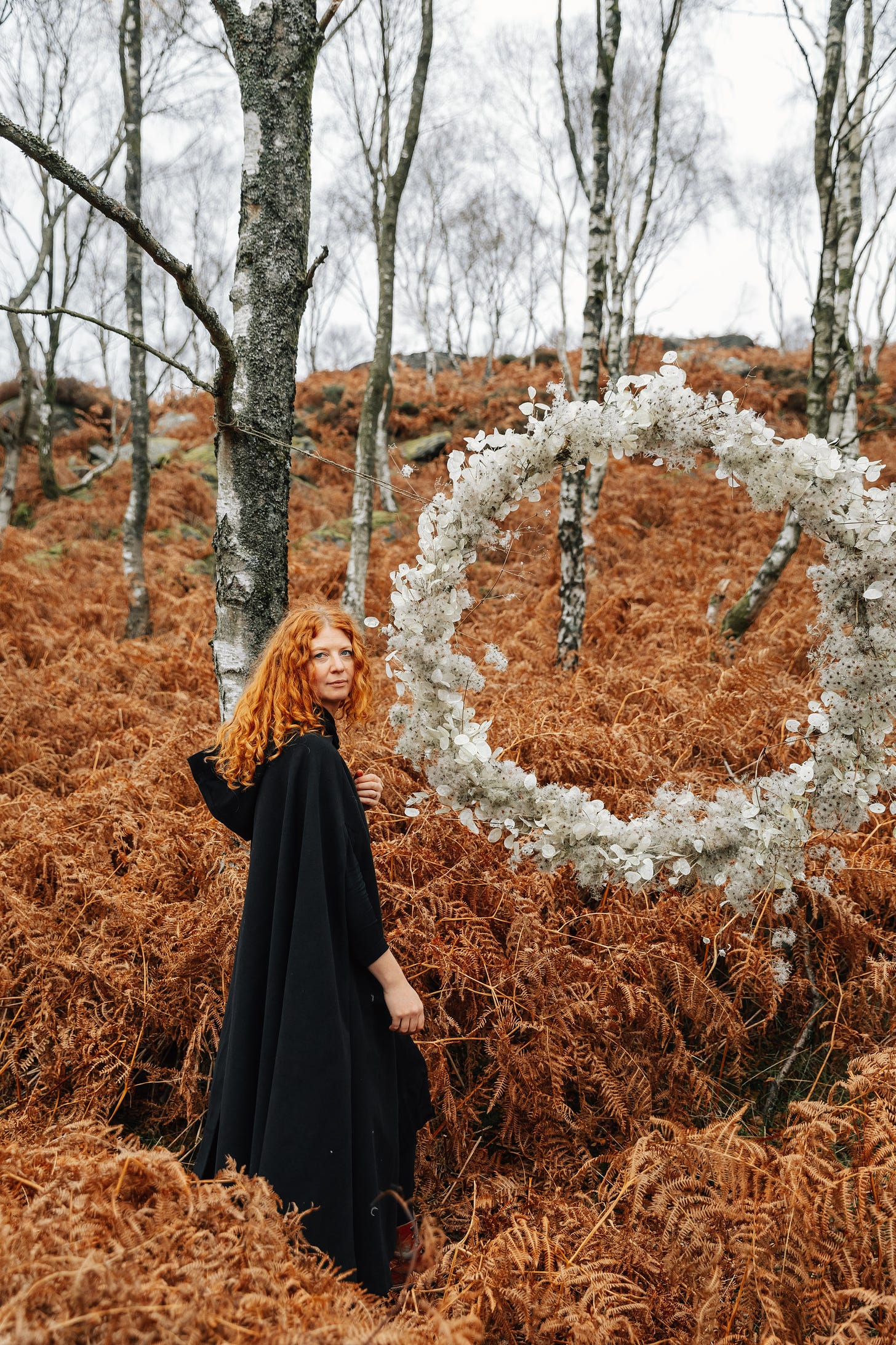 In the wintery woods, a giant hoop of honesty and winter foliage. A woman with hair that looks like the bracken stands about to step into the hoop, looking back at the viewer. She is wearing a dark cloak and boots.