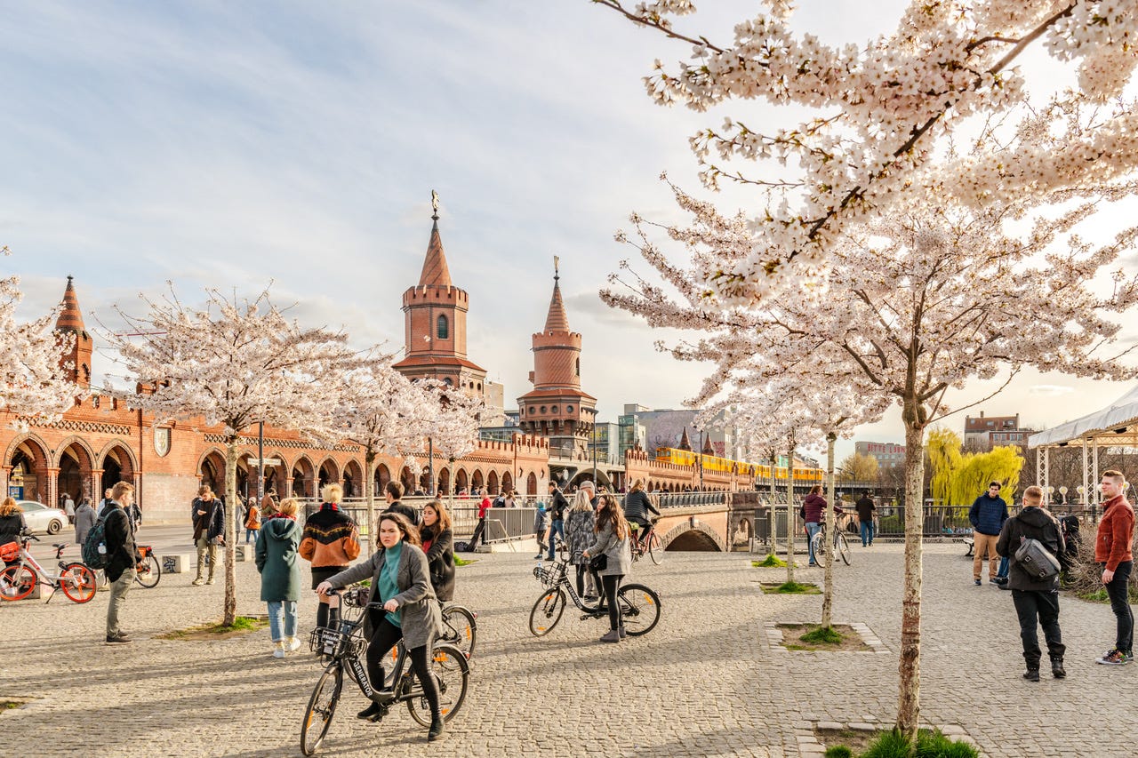 Cyclists and people walking in a cobbled area with blossoming trees in front of the Oberbaum Bridge.