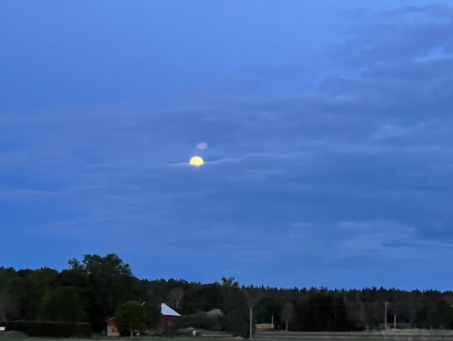 The full moon peeping through a cloudy sky hanging over a strip of forest and farmland