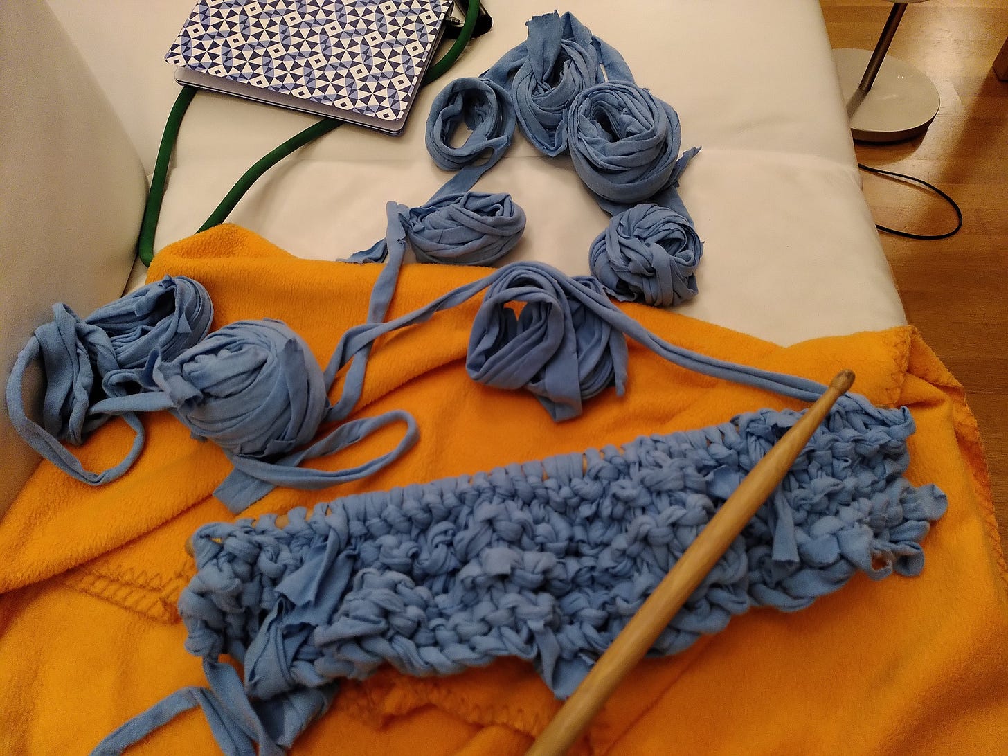 knitting work in progress, light blue piece, drumstick "needle" laying across piece, several balls of jersey fabric yarn next to piece set against orange blanket background.