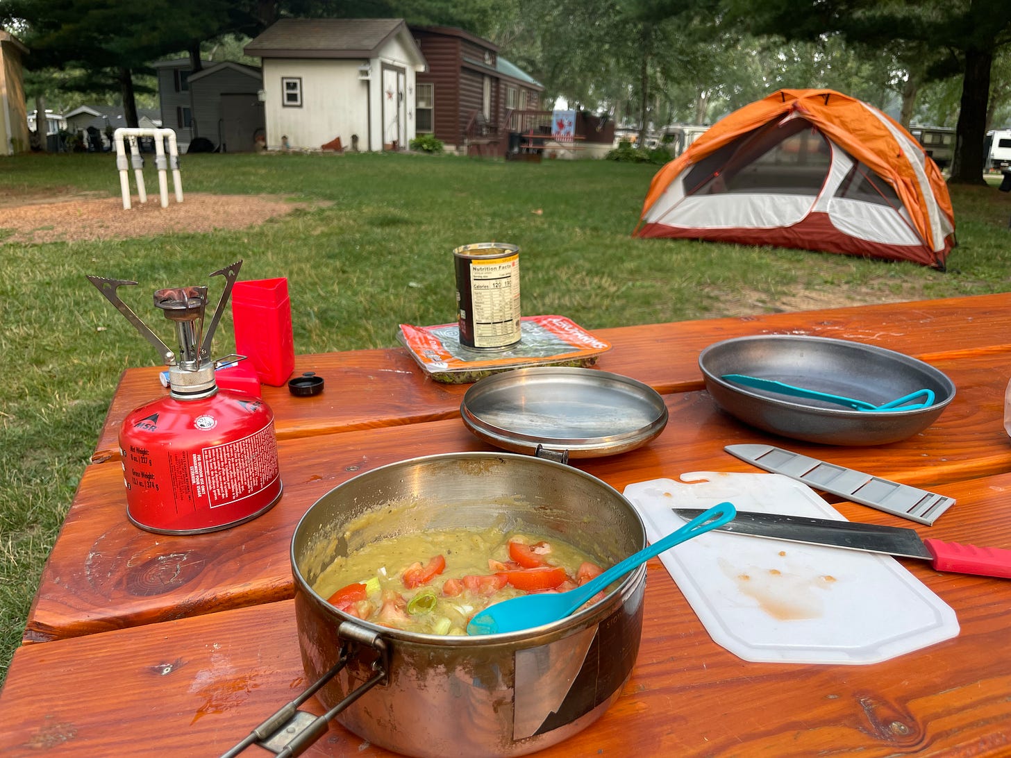 Photograph of a meal being prepared on a picnic table. There's a camp stove, a pot with green soup, a plate, a knife and cutting board.