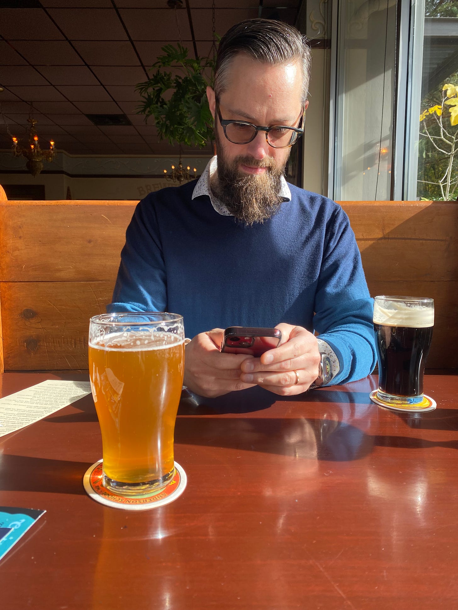 Jeff at McMenamin's, wearing a blue sweater and sitting in a wooden booth. On the table, sun shining in, are a glass of IPA and a glass of stout.