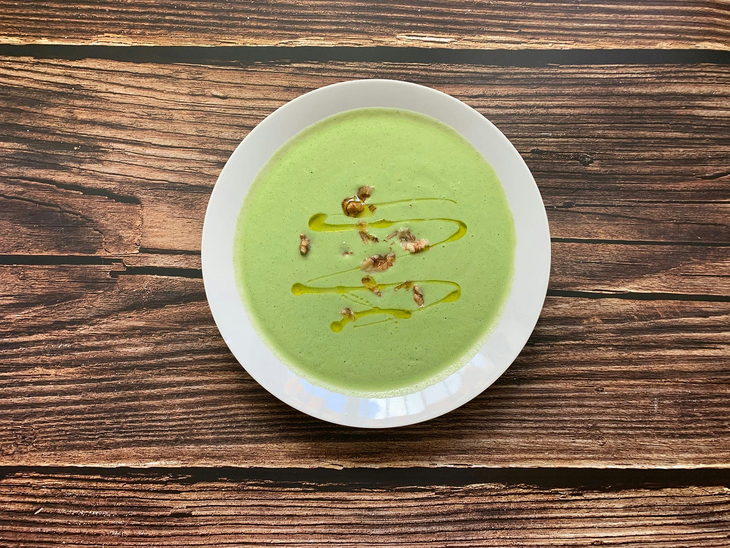 White bowl filled with green pea soup with walnuts and a little oil garnish, on a rough wooden background