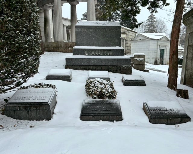 The Wollman family plot: six headstones covered in snow before a monument engraved "Wollman." In the row closer to the camera in the center is Benjamin's grave with a shrubbery behind it. To the left is Kate's grave, by a tree with a partial shrubbery behind the headstone.