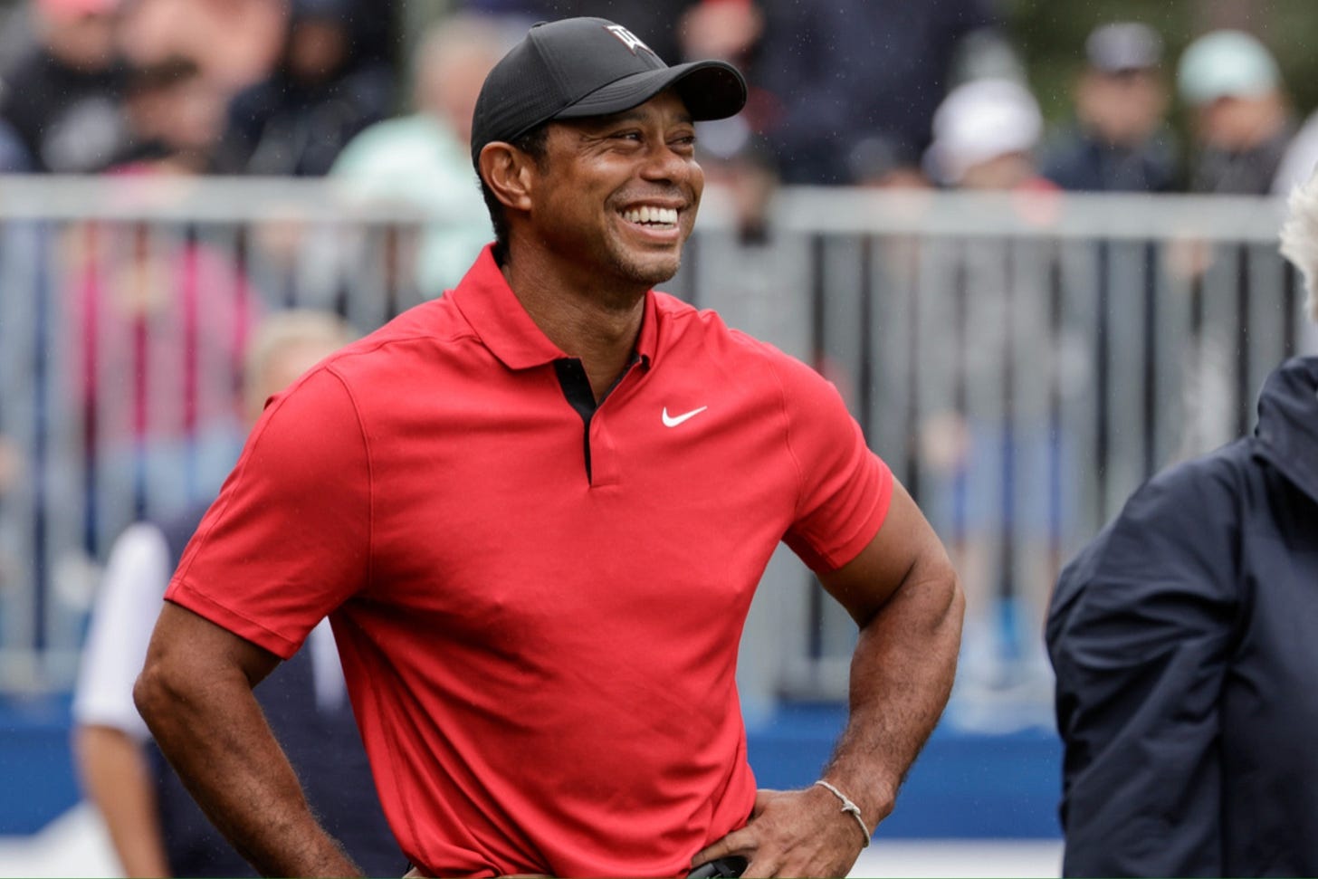 Tiger Woods smiling broadly while wearing a red Nike polo shirt and black cap at a golf tournament.