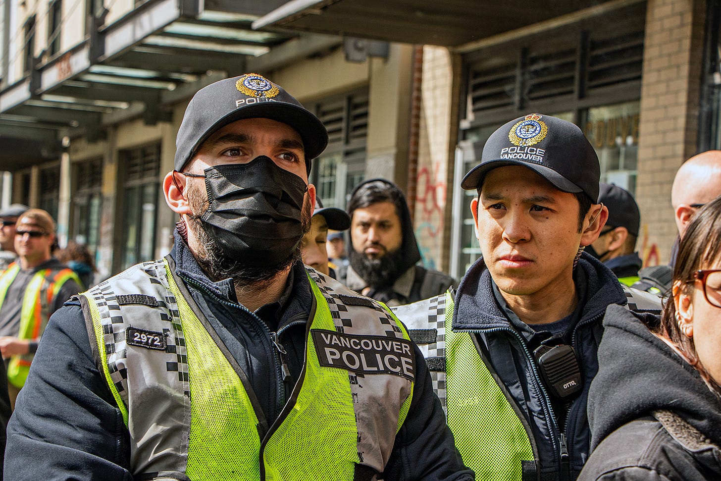 A police officer stands looking ahead. he is wearing a mask, a hat that says "police" on it and a yellow vest. another police officer, also in a hat and vest, stands to his left (our right) and just behind him, also looking forward and kind of to our left/his right.