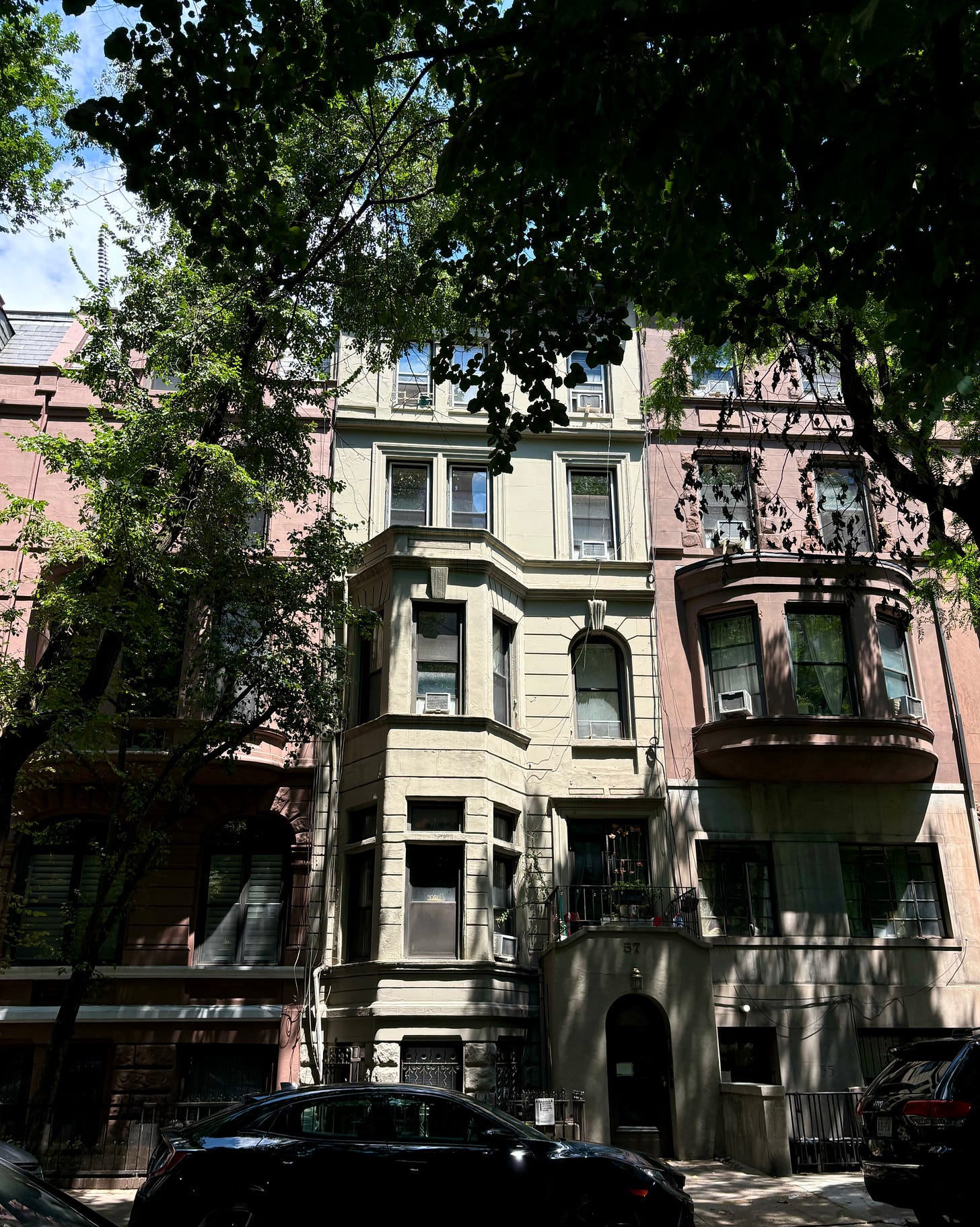 A light stone house between two blush townhouses. It is framed by leafy trees and dappled in shadow.