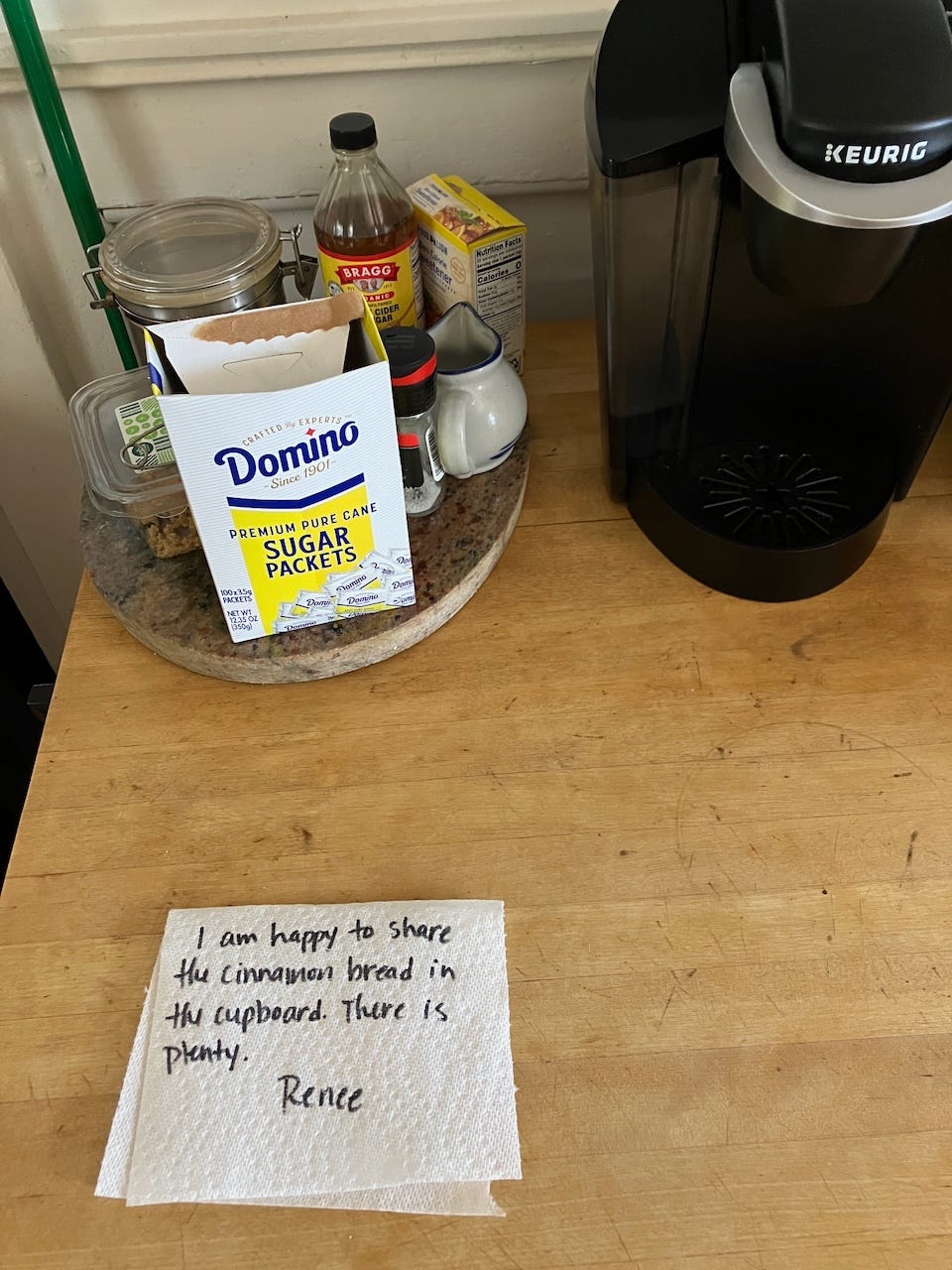 Coffee maker next to box of sugar packets and a note written on a paper towel reading "I am happy to share the cinnamon bread in the cupboard. There is plenty--Renee"