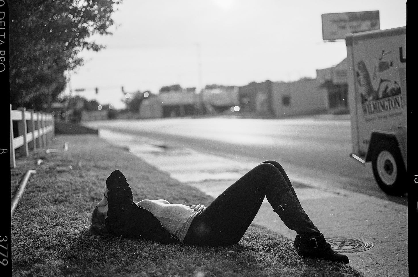 A black and white film photo. Amber is lying on a narrow, grassy patch next to a road. Her knees are bent. One hand rests on her stomach. Her other hand is on her forehead, obstructing the view of her face.