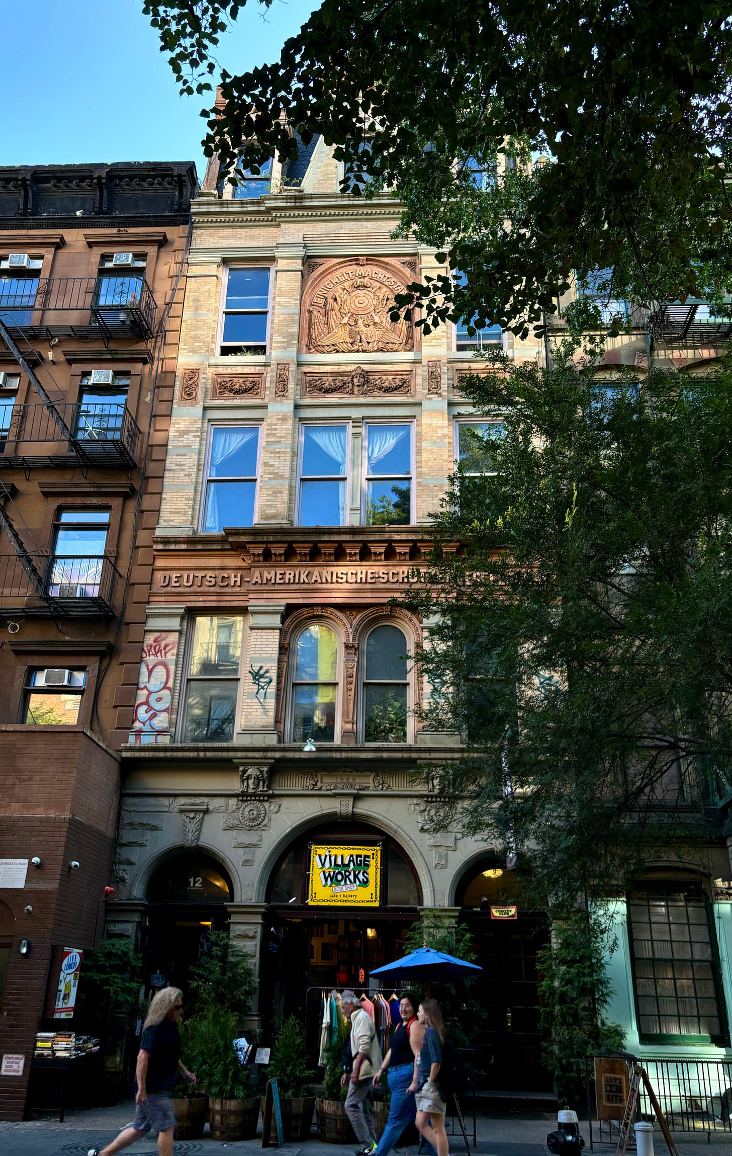 A full view of the light brick building engraved German-American. There is a yellow sign that reads VILLAGE WORKS Book Shop in black letters..