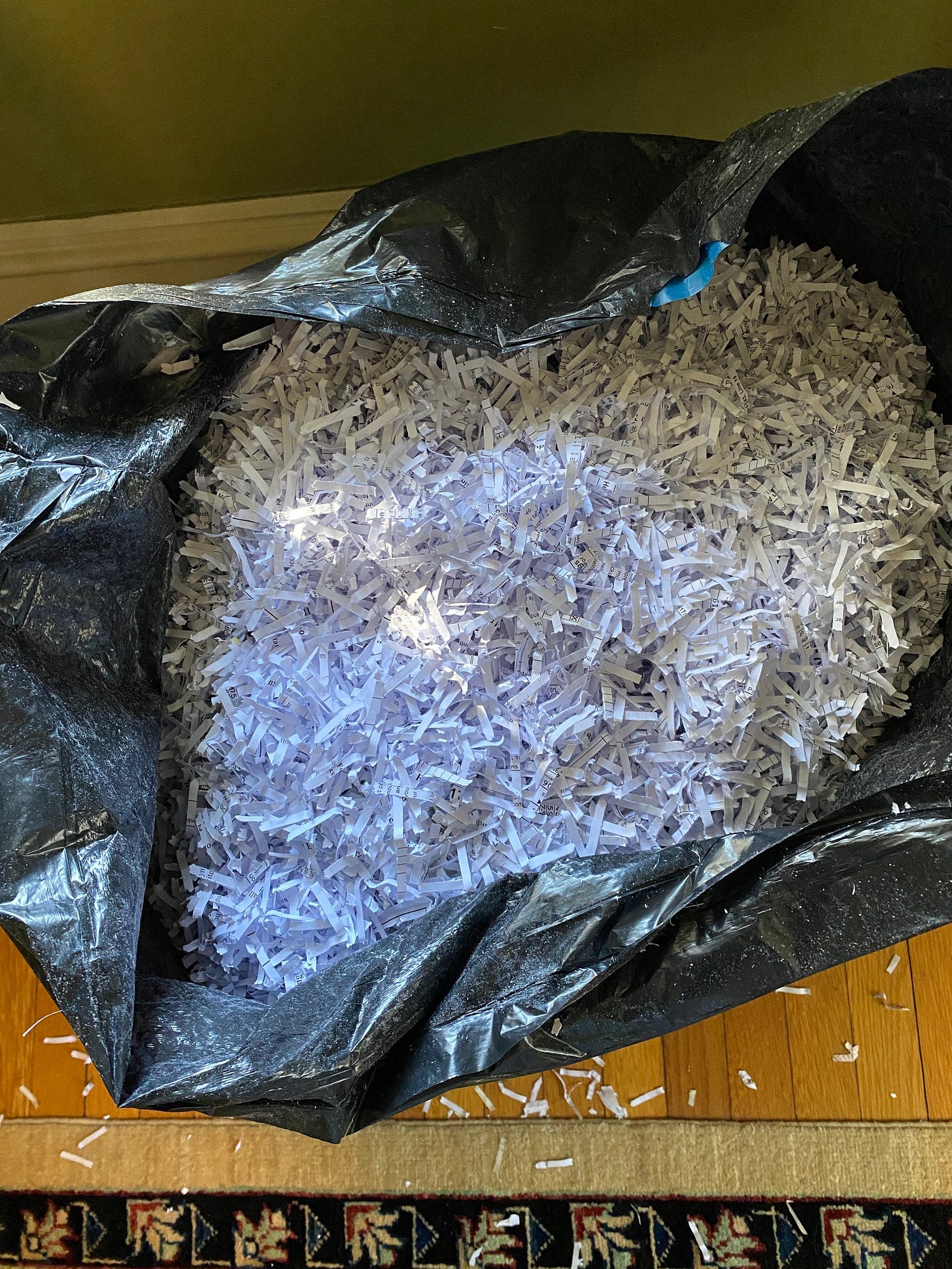 View from above, looking into an open black plastic trashbag filled with shredded white paper, sitting on a hardwood floor.