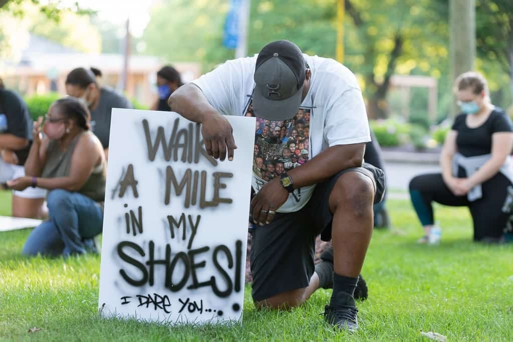 Kneeling man with "Walk a mile in my shoes!" sign.