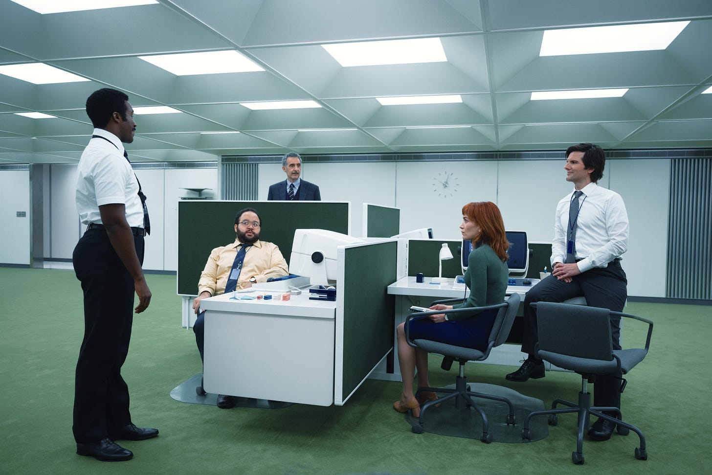 Still image from Severance, showing five officeworkers in a sparse, 70s-style office. The computer monitors are so deep they take up half the desk. Green partitions separate the four desks.