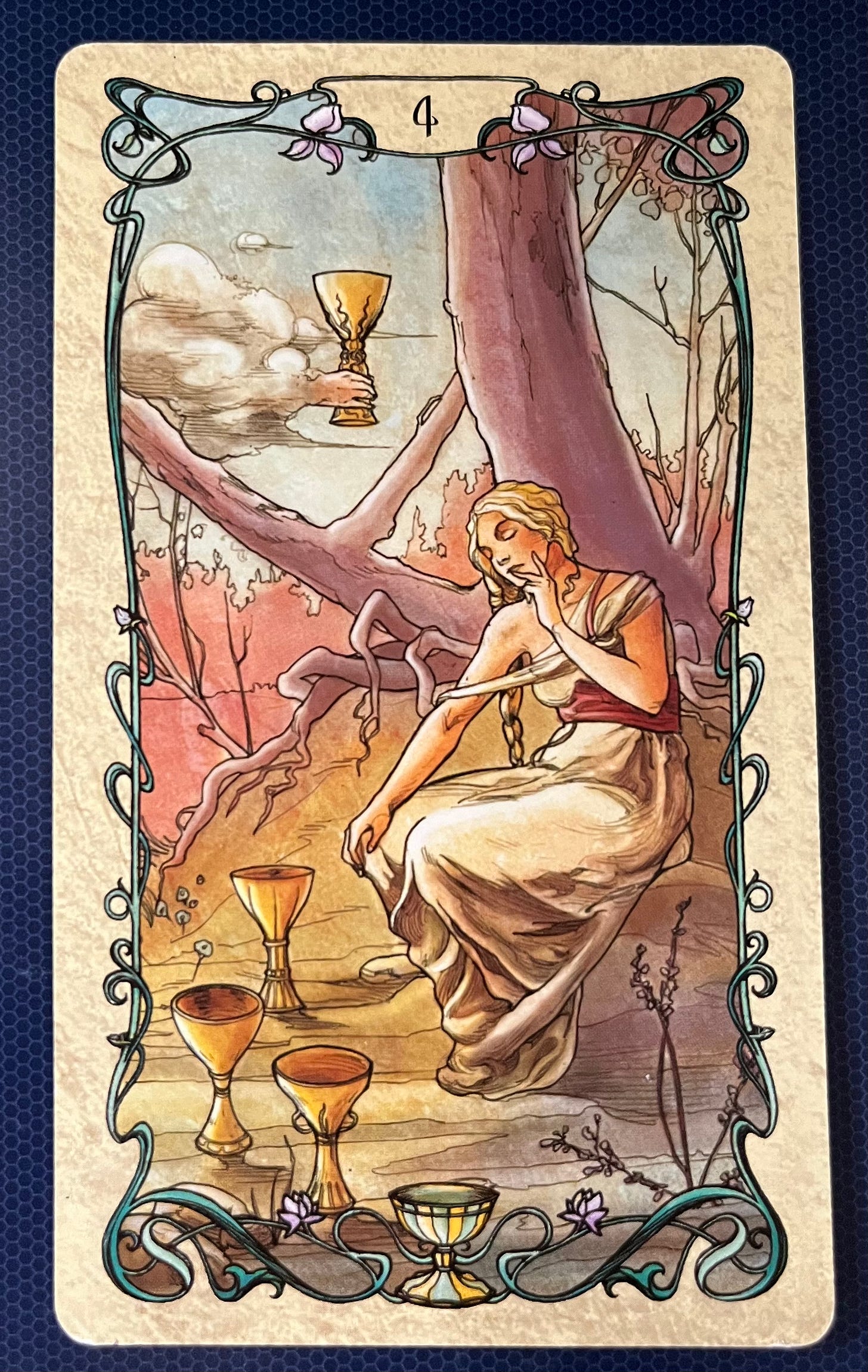 The image is of a tarot card specifically the four of cups with an ornate border and the number 4 at the top. The art is in the style of Mucha. It depicts a person sitting under a tree, appearing to be in deep thought or contemplation. The individual is wearing classical attire and holding their head with one hand. There are four cups around the individual; three on the ground and one being offered by a clouded hand emerging from the sky. The background shows bare trees and clouds, giving an autumnal or barren feel to the scene. The intricate border features vine-like decorations with small flowers interspersed throughout. 