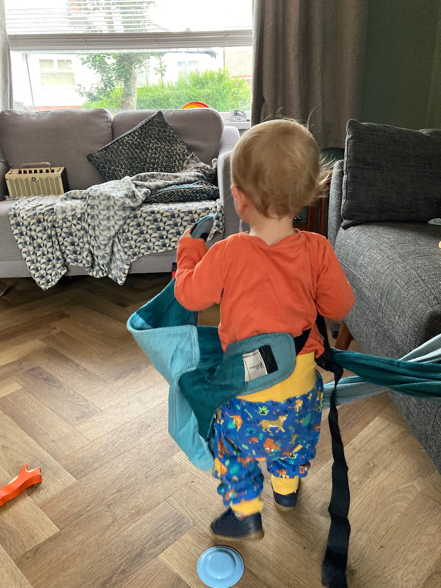 A blonde todller is facing away from the camera. He is holding a turquoise woven baby carrier and has wrapped it round his body