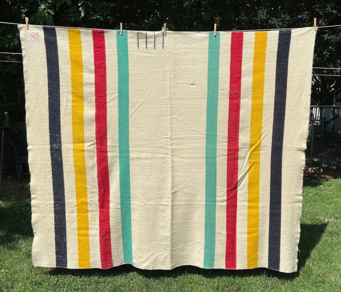 May include: A white wool blanket with black, yellow, red, and teal stripes. The blanket is hanging on a clothesline outdoors.