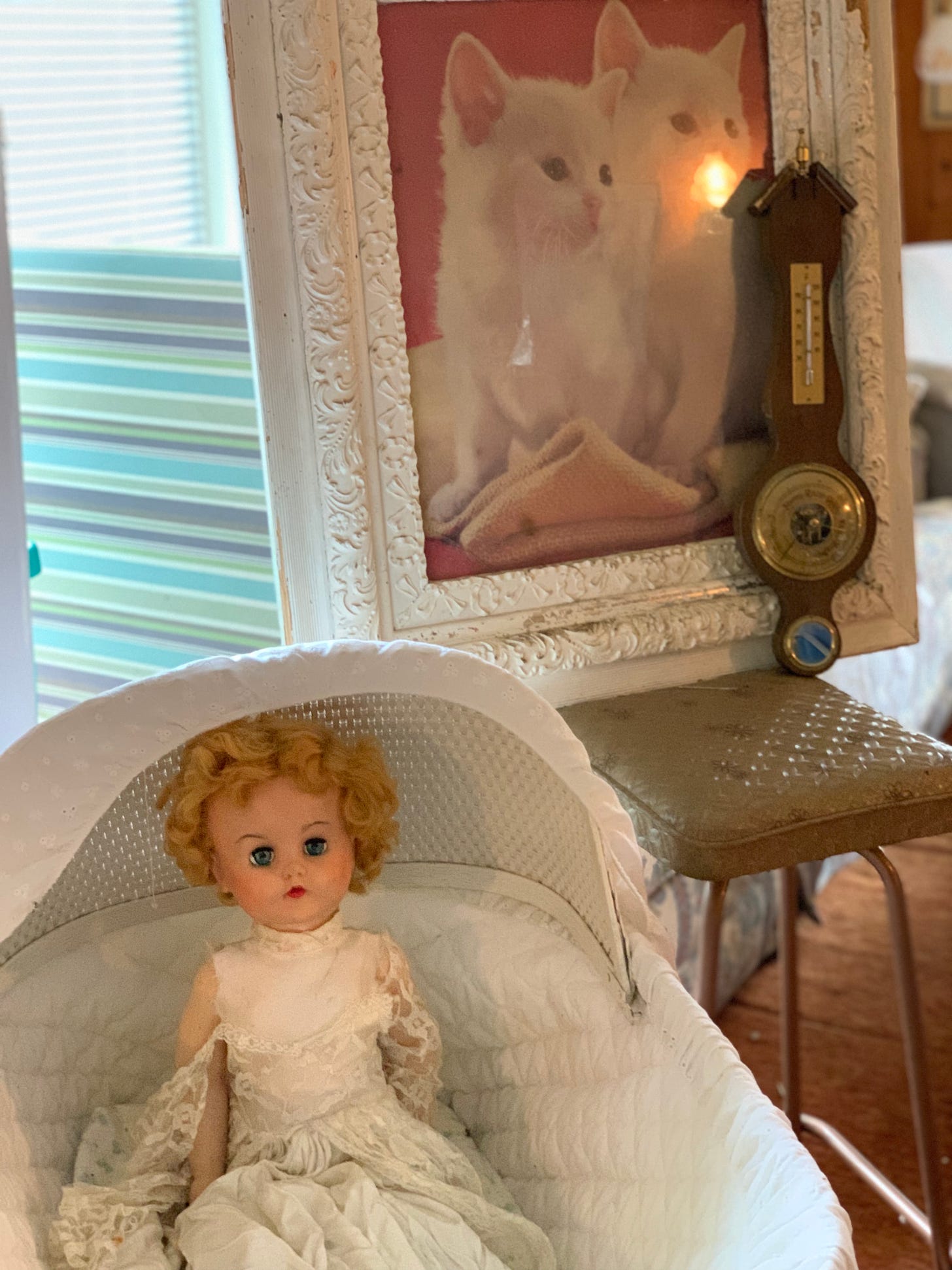 A doll, with blonde hair and dressed in white sitting in a white bassinet. Behind her is a white picture frame featuring a picture of a white cat. 