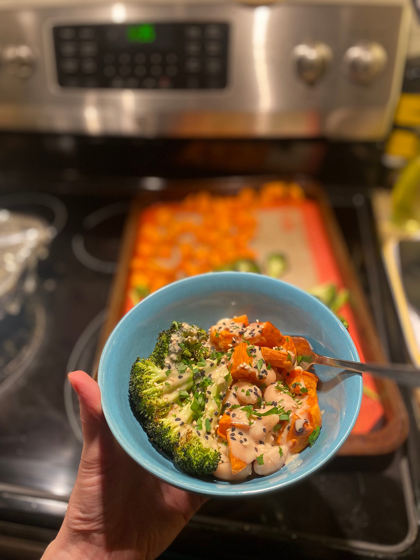 A blue bowl of roasted orange sweet potato and broccoli florets with beans and brown rice, topped with black and white sesame seeds and parsley. In the background on top of the stove is a sheet pan with the remaining roasted vegetables.