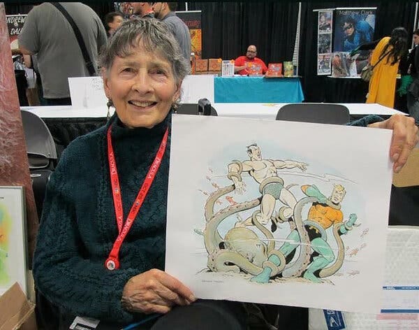 Ramona Fradon, a gray-haired woman wearing a dark sweater with an ID badge on a lanyard, holds up a drawing of two superheroes battling an octopus.