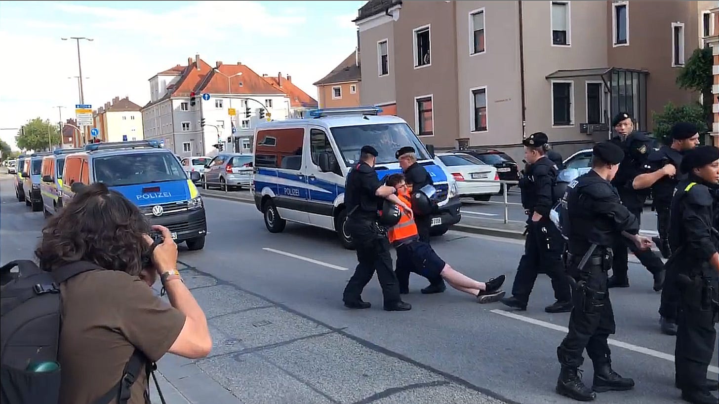 Bild 2: Polizei nimmt einen der Protestteilnehmer in Gewahrsam und trägt ihn aus der Versammlung heraus zum Polizeitransporter. Ein Fotograf hält die Szene auf Kamera fest.