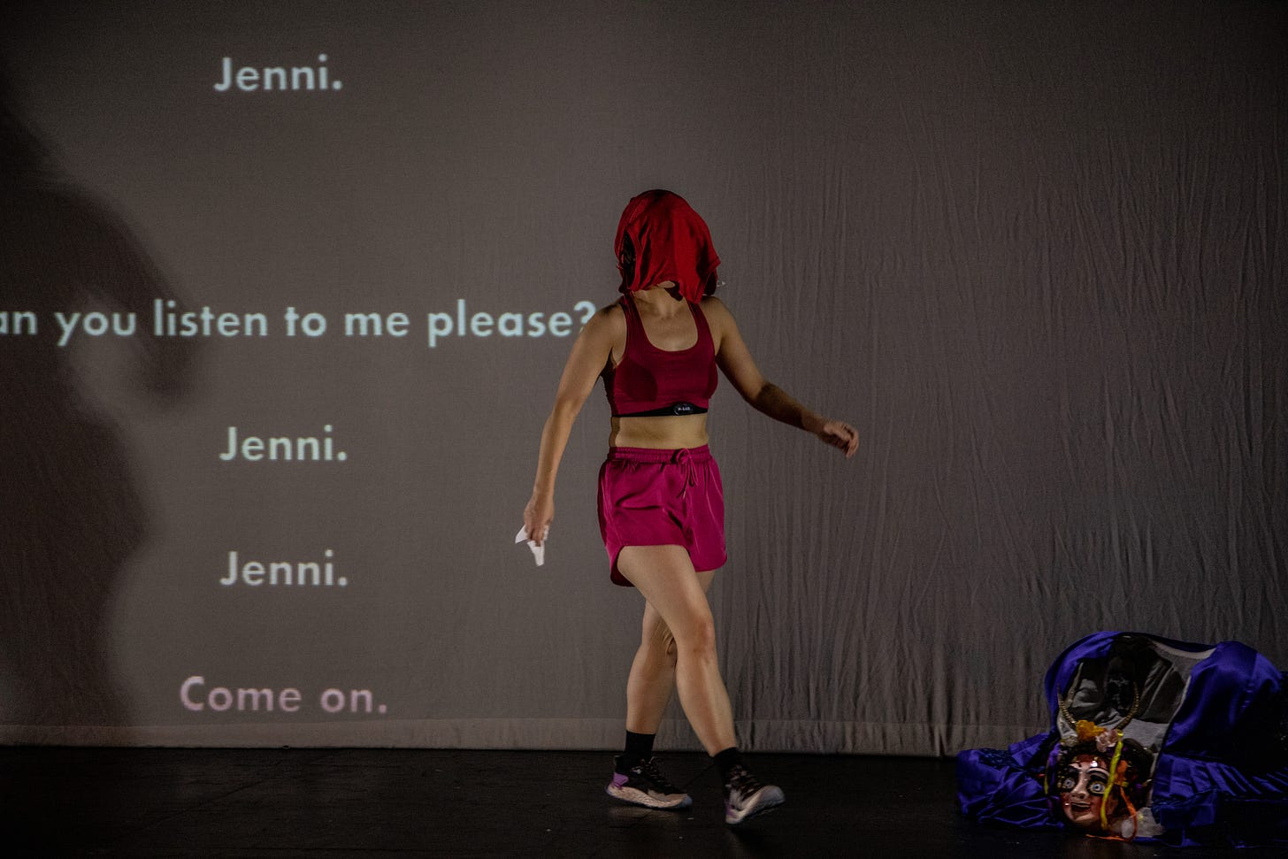 A photograph of Jennifer Jackson in Endurance, with a running top over her head in front of projected text reading 'Jenni. Can you listen to me please? Jenni. Jenni. Come on.'