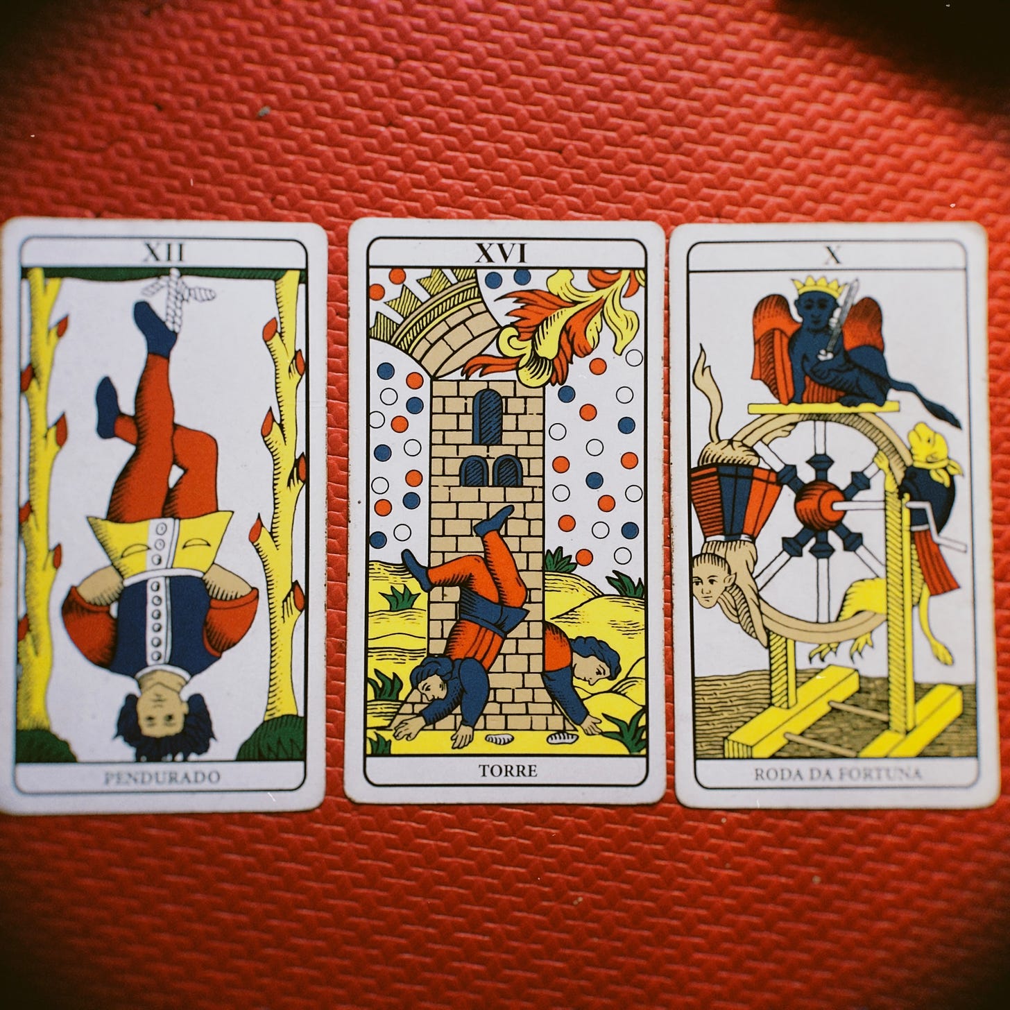 Três cartas do tarot de Marselha em cima de um tapete vermelho de Yoga: Um homem pendurado pelo pé; dois homens caindo de uma torre em chamas; a roda da fortuna com animais que sobem e descem.
