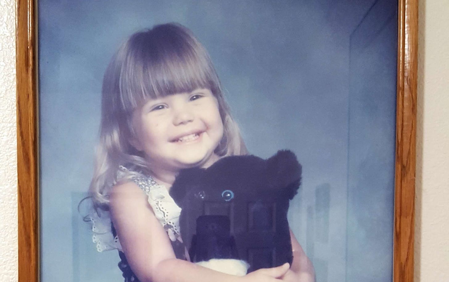 A blond three-year-old girl in a blue dress hugs a black teddy bear
