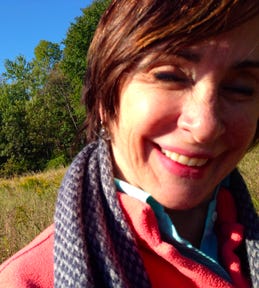 A close-up photo of a woman smiling with trees and grass in the background. She is wearing a patterned scarf, a pink jacket and a blue button up shirt.