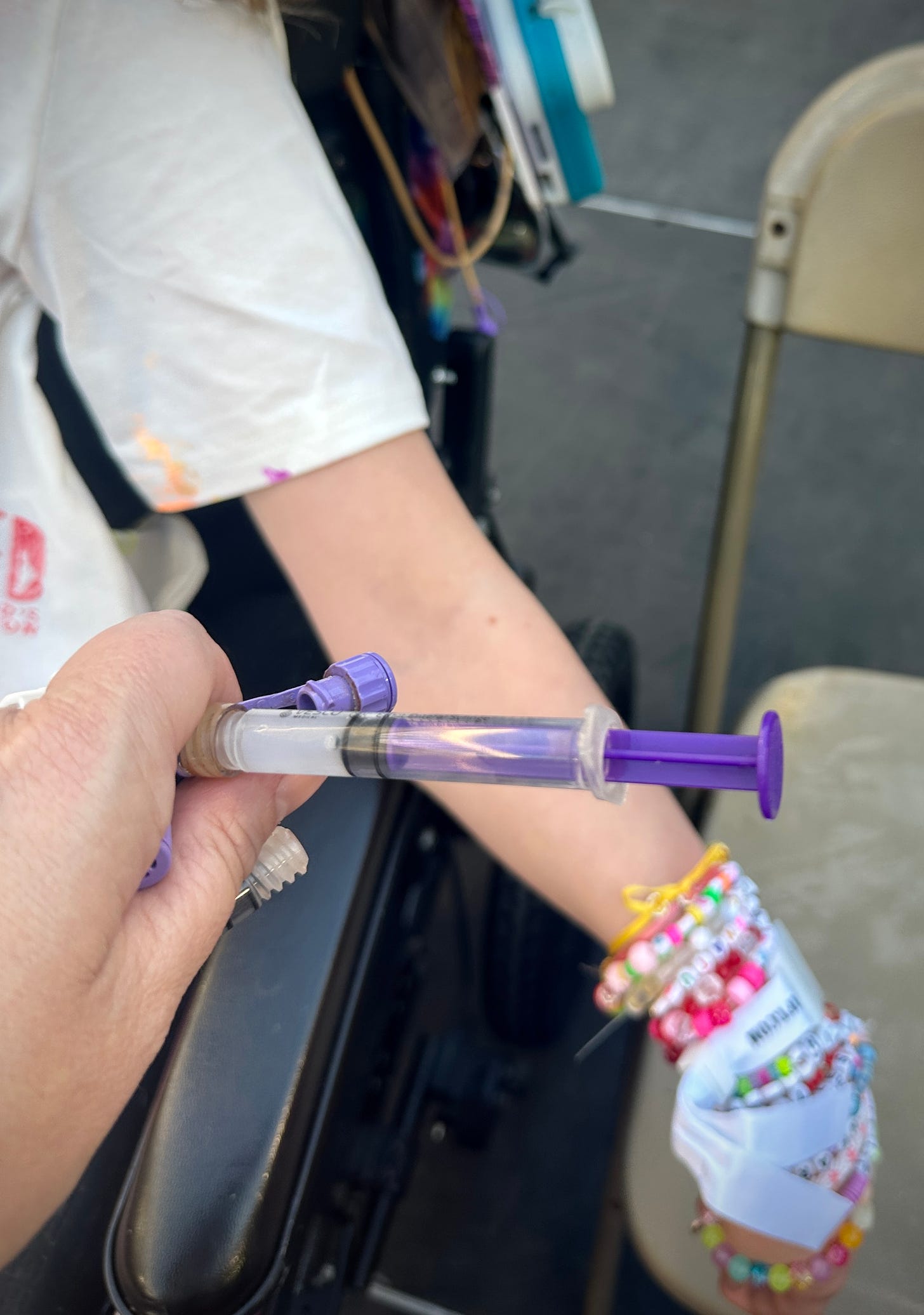 A closeup of a mother’s hands pushing a purple syringe of medication into a purple tube set. In the background, a young girl’s arm hangs, covered in beaded bracelets. They are on a platform with a folding chair nearby. 