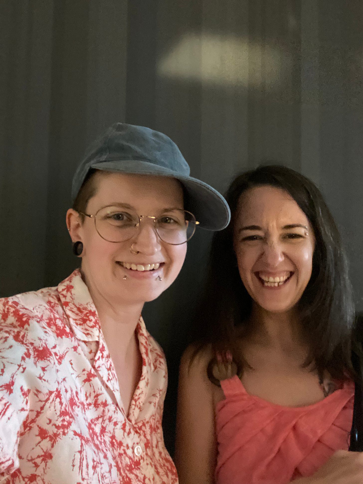 a photo of me and Steph in front of a grey background. I am wearing my gold-rimmed glasses, a denim blue baseball hat, a white shirt with a camp collar and an abstract dark pink pattern. Steph is wearing a pink sundress with crossover detail in front. Her dark hair is loose and hanging over her shoulders to the neckline of the dress. We are both smiling, as if we've just been laughing.