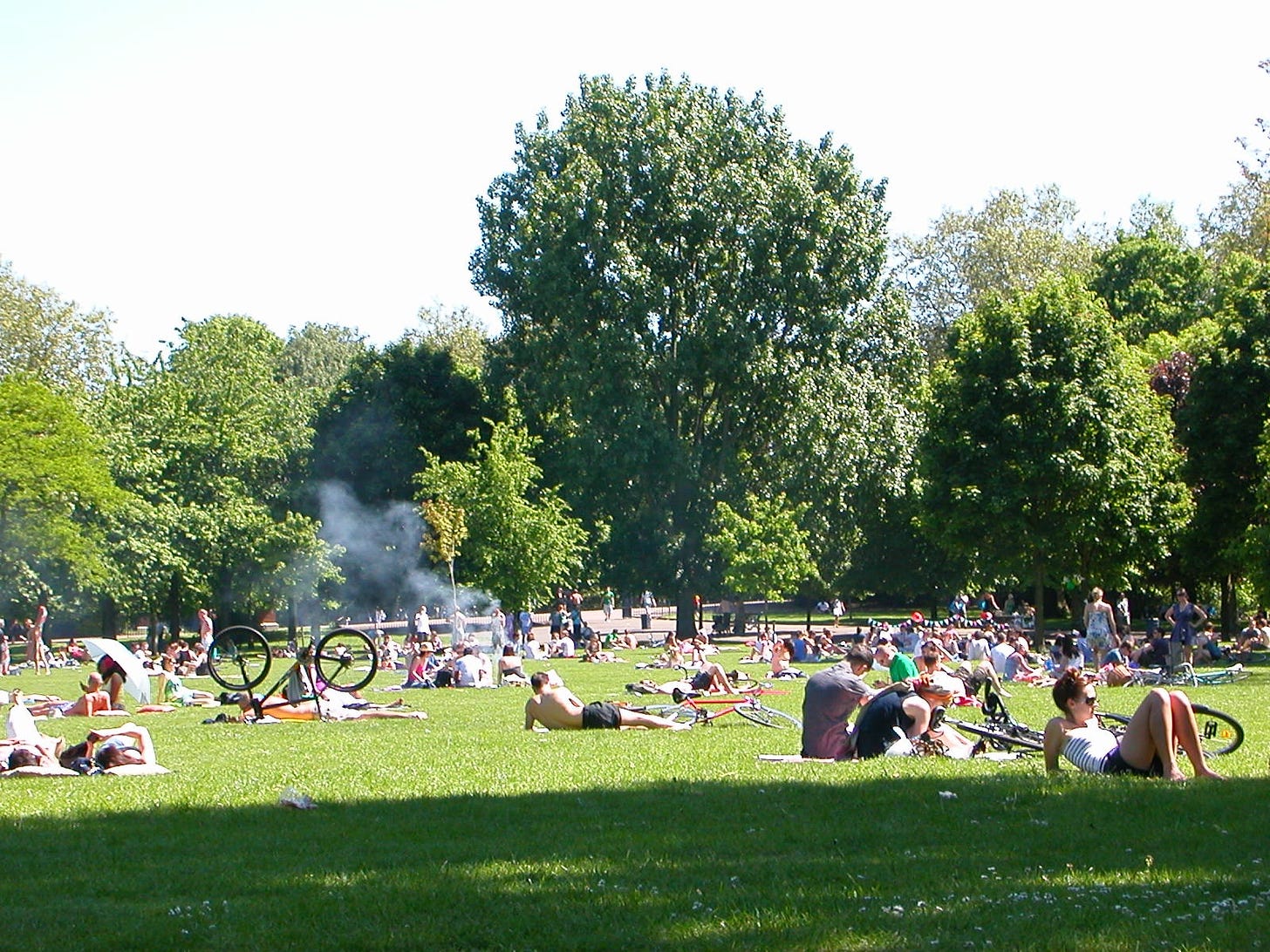 Victoria Park in London, during a warm summer day, with the grass covered with people sunbathing, chatting, kicking a ball and even someone BBQing something.