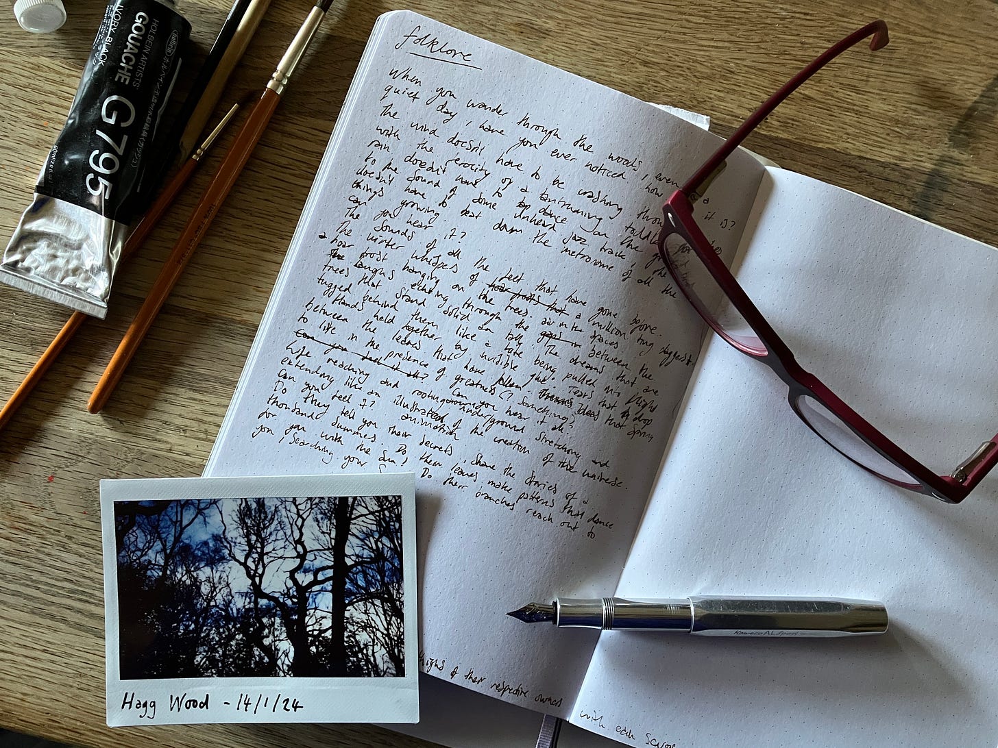Notebook with the written post in sits atop a wooden table. Red glasses and an aluminium fountain pen sit atop the open notebook with an Instax photo of woodlands.