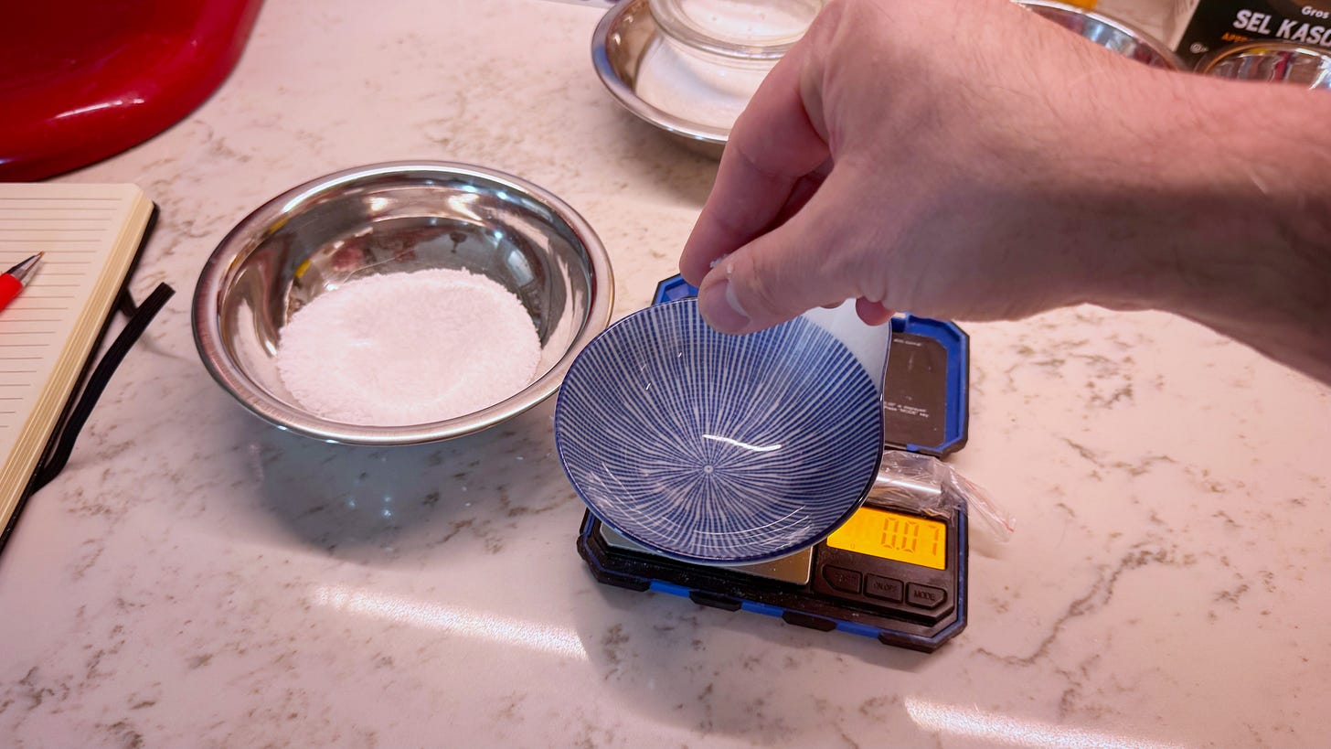 A hand holding a pinch of salt, about to drop it into a ceramic bowl sitting on a digital jeweller's scale Beside the scale is a small, stainless steel bowl full of salt. 