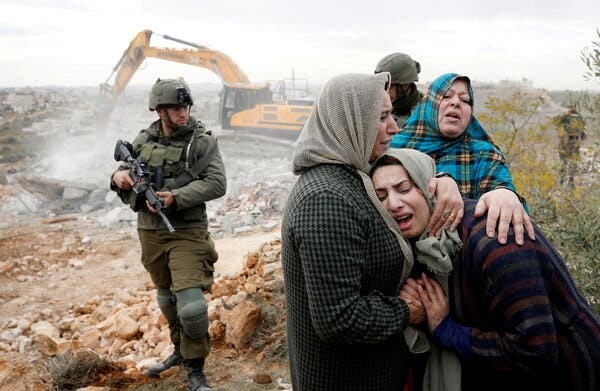 An armed soldier stands near a group of crying women. There is a bulldozer in the background.